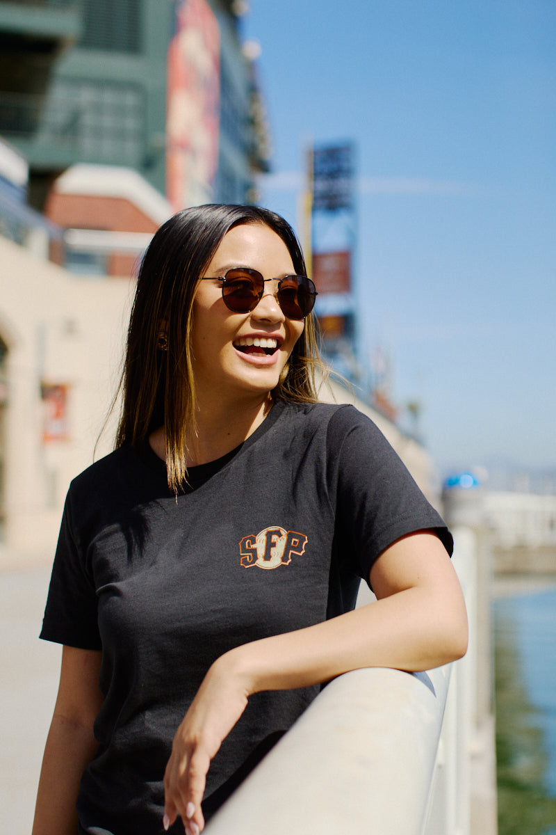 A smiling woman in sunglasses leans on a railing outdoors, wearing the Psychos Logo Tee—a hand-printed black SFP t-shirt from San Francisco.