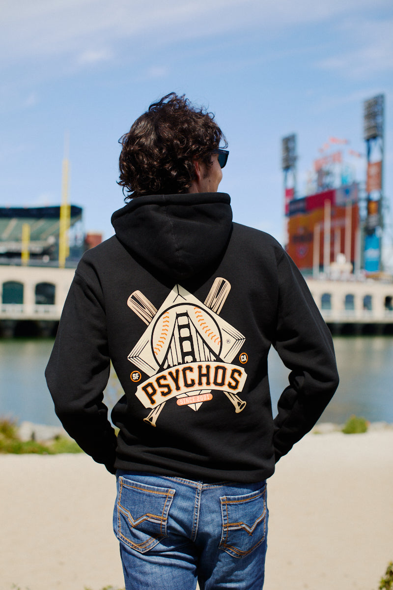 Wearing a Psychos Logo Pullover Hoodie, someone stands by a serene San Francisco waterway. This unisex pullover boasts baseball graphics and Psychos text, blending style and comfort perfectly with the coastal scenery.