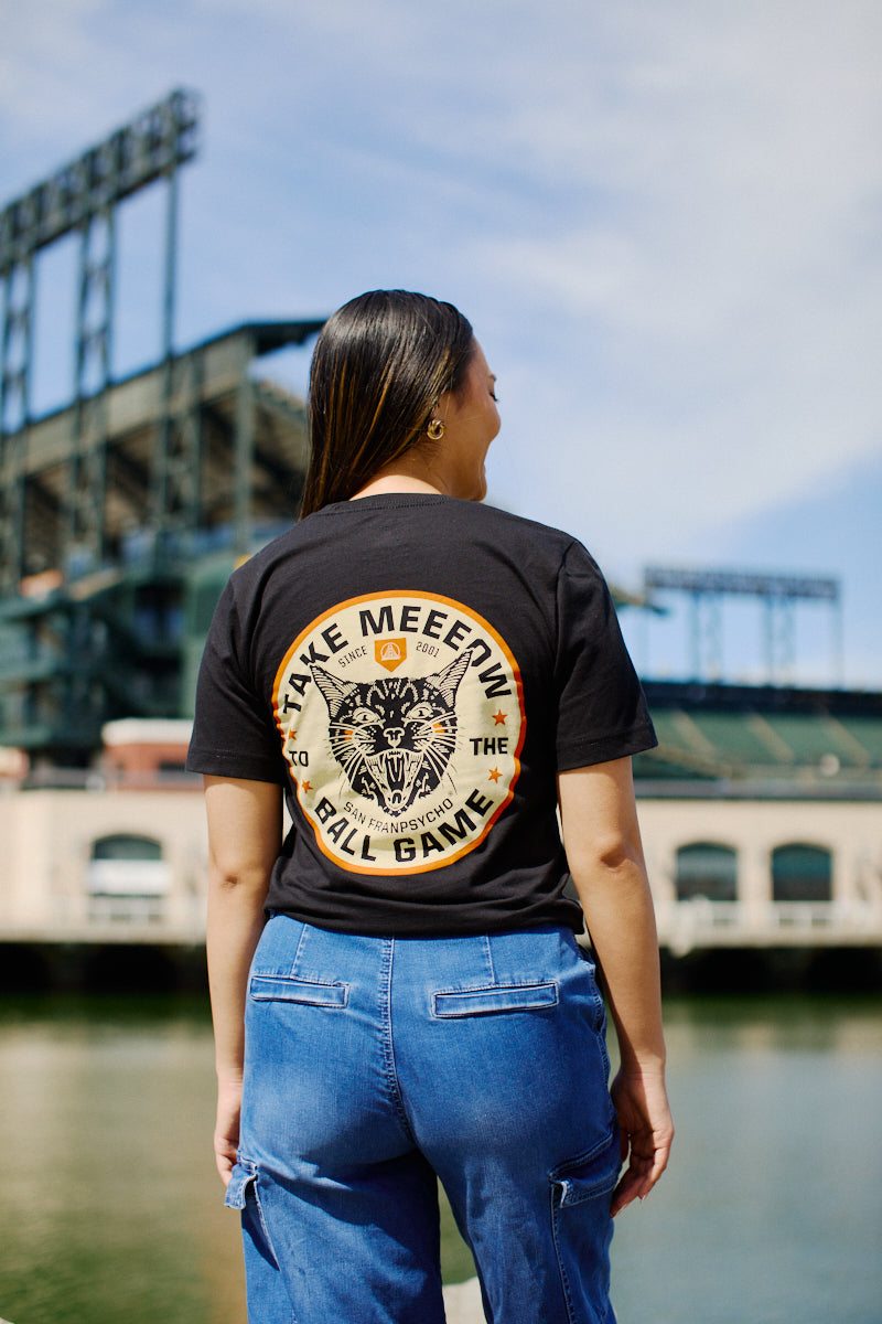 A woman confidently stands by the stadium and water, proudly wearing the Black Take Meeeow Tee with a hand-printed animal graphic on the back, radiating black and orange pride.