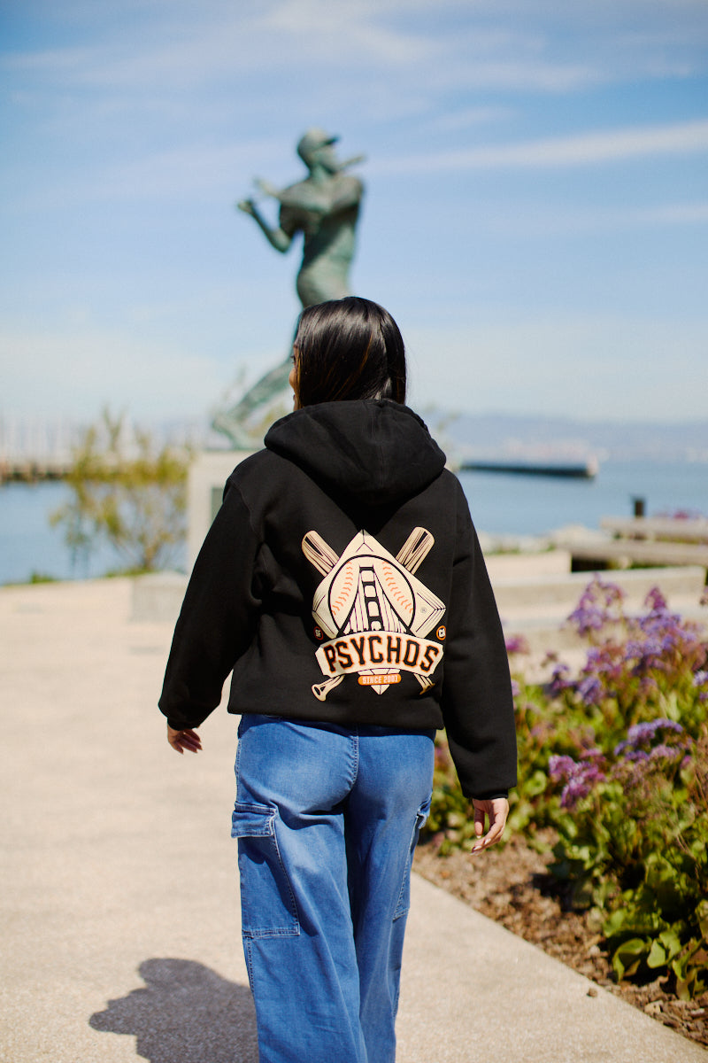 A person wearing a Psychos Logo Pullover Hoodie and jeans strolls along a waterside path with a statue in the background, perhaps savoring the cool San Francisco breeze.