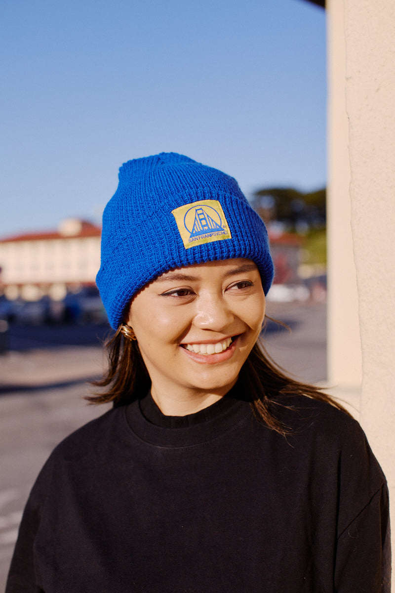 Smiling woman outdoors on a sunny day wears a Royal Blue Waffle Beanie with Yellow/Blue Logo and a black shirt.