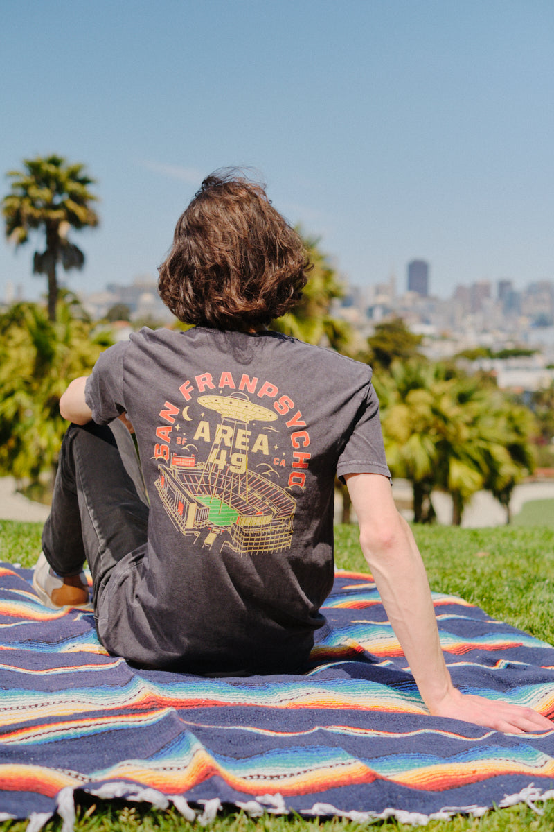 Relaxing in the park on a colorful blanket, someone wears the Area 49 Tee—a vintage style “San Francisco Bay Area” shirt crafted from 100% ring spun cotton—with the city skyline in view.