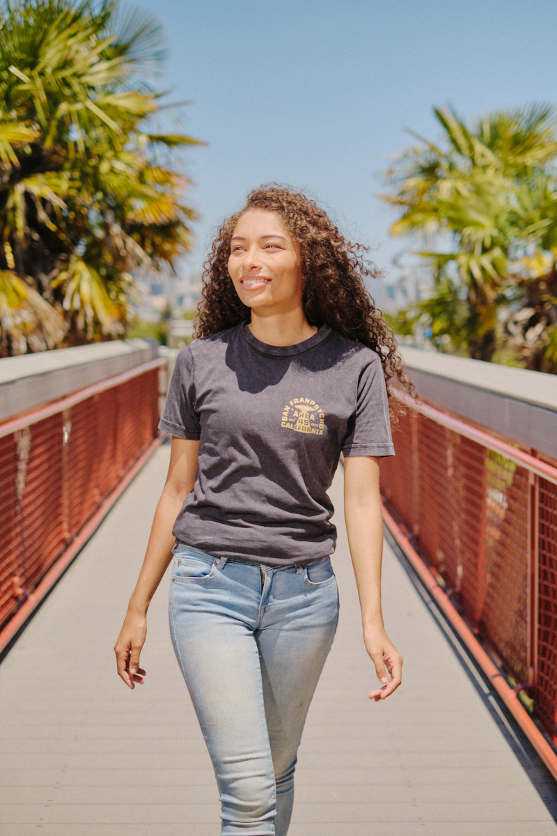 A woman with curly hair smiles as she walks on a sunny bridge lined with palm trees, wearing jeans and the Area 49 Tee.