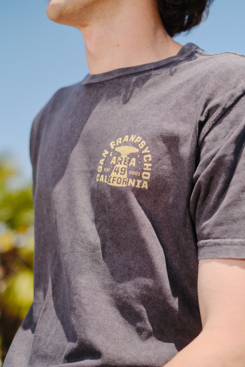 Under a blue sky, a person wears the Area 49 Tee in faded black, featuring San Francisco Area 49 California text. This vintage-style shirt delivers laid-back vibes, perfect for sunny days outdoors.