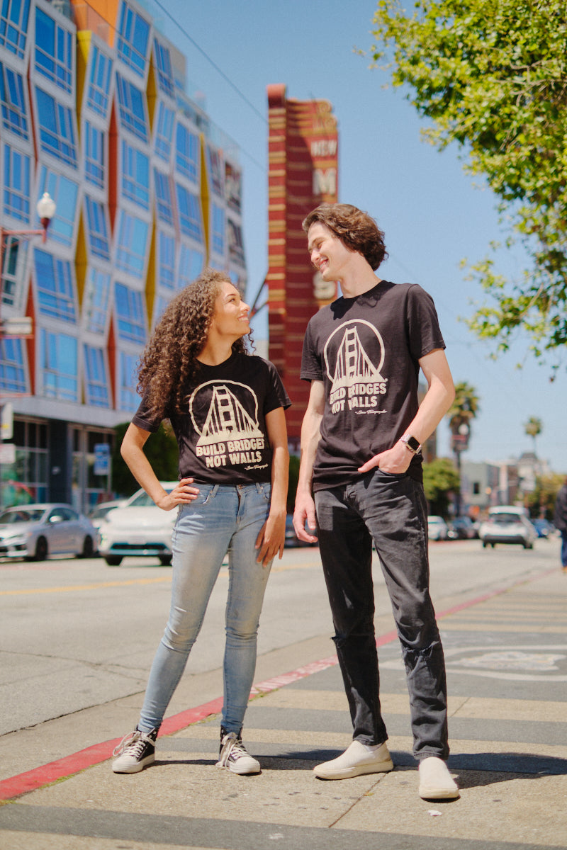 Two people stand on a city sidewalk, smiling at each other in matching Build Bridges Not Walls Tees.