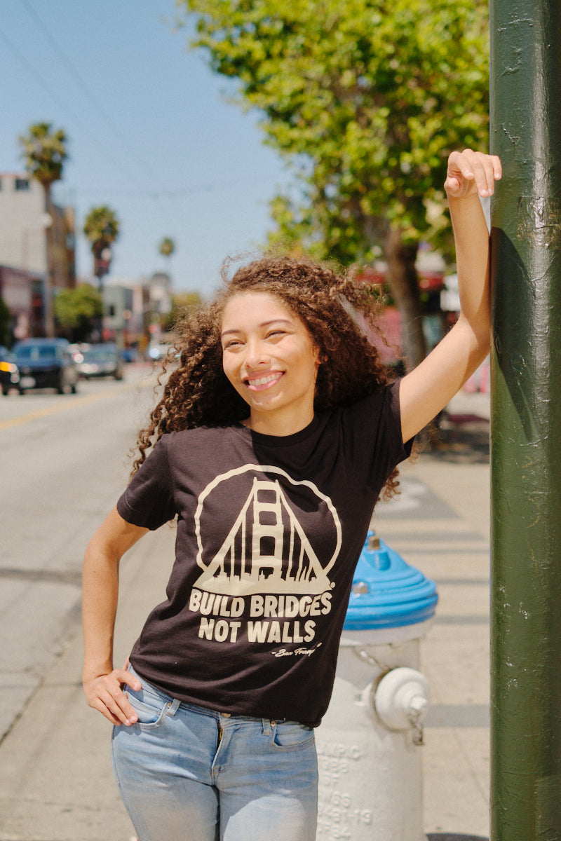 A smiling woman stands by a pole on a sunny city street, wearing the Build Bridges Not Walls Tee.