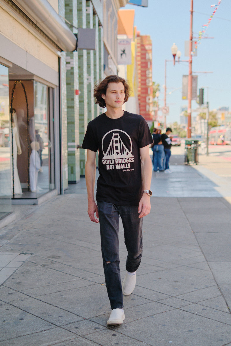 A man walks on a sunny city sidewalk wearing the Build Bridges Not Walls Tee, made from 100% airlume cotton.