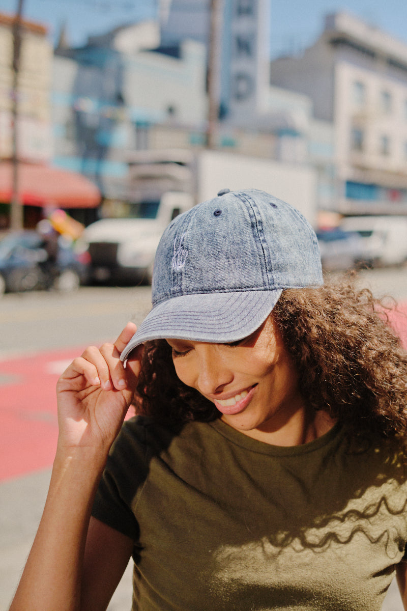 A woman smiles outdoors in front of city buildings, wearing a green shirt and the Blue Denim Peace Bones Dad Hat featuring an embroidered design.