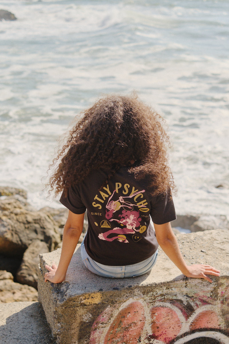 A person with curly hair sits on a rock by the ocean, wearing the Psycho Siren Tee—crafted from soft Airlume cotton and featuring a colorful, hand-printed design.