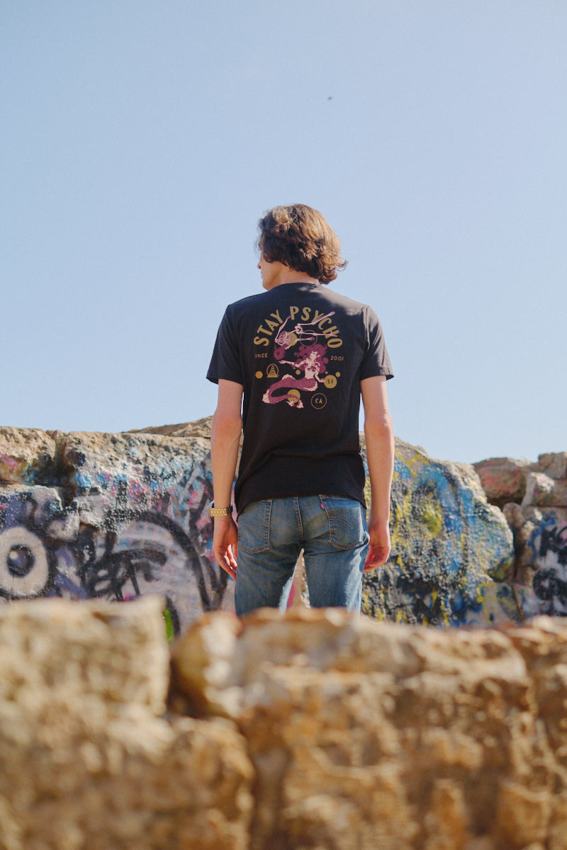 Wearing the Psycho Siren Tee, made from soft Airlume cotton and hand printed, a person in jeans stands on rocks before a graffitied wall under a clear sky.