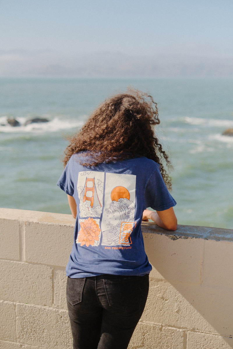 A person with curly hair wearing the Sunset Dahlia Tee, inspired by Japanese art with a blue graphic design, stands by a wall and looks out over the ocean on a sunny day.