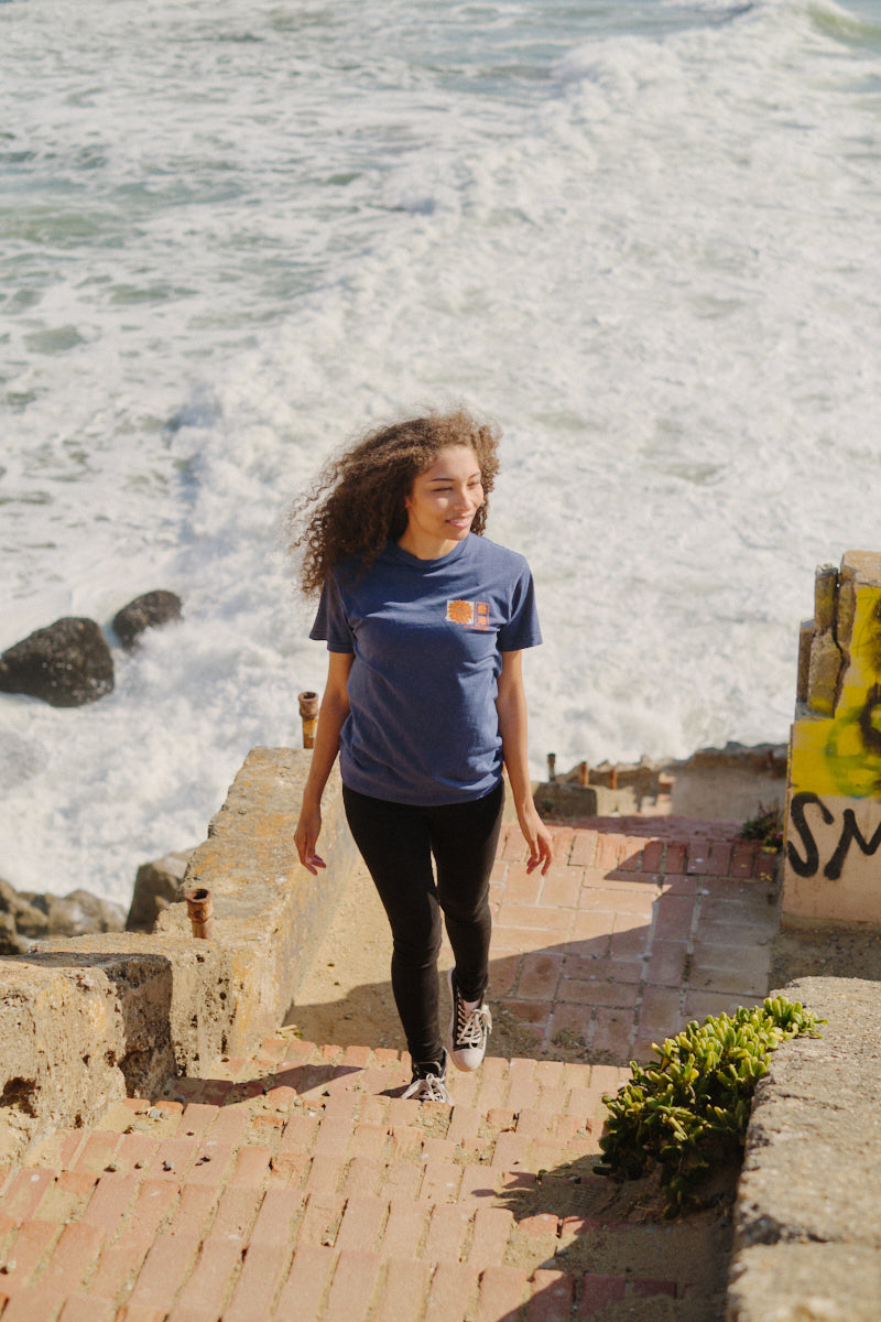 A woman with curly hair, smiling as she walks up seaside steps on a sunny day, wears the Sunset Dahlia Tee with waves in the background.