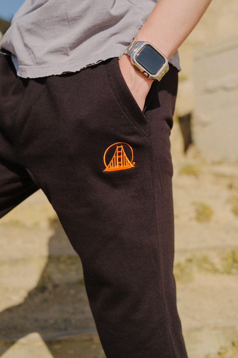 A person wearing Black & Orange Embroidered Logo Joggers featuring an orange Golden Gate Bridge logo, with a hand in the pocket and a smartwatch.