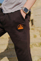 A person wearing Black & Orange Embroidered Logo Joggers featuring an orange Golden Gate Bridge logo, with a hand in the pocket and a smartwatch.