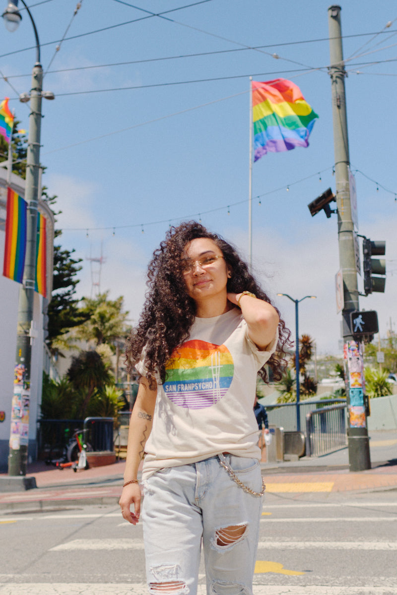 A person with curly hair wears the Rainbow Sutro Tee, standing outside with pride flags in the background.