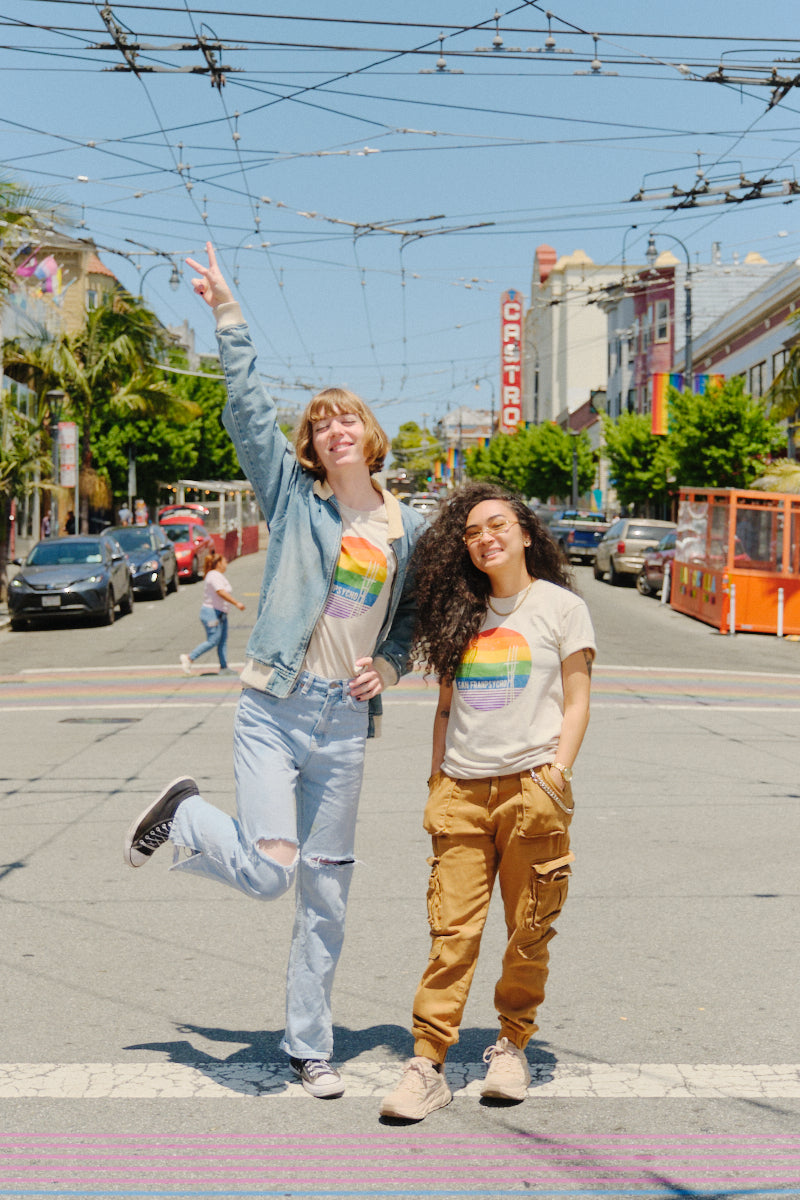 Two people in Rainbow Sutro Tees celebrate pride and unisex style near the SF LGBT Center, posing on a city street under blue skies—one jumping, one smiling.