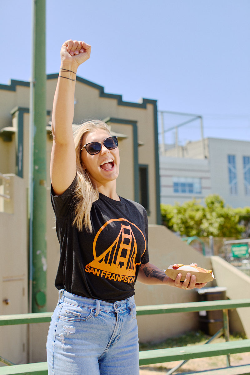 Smiling woman in sunglasses raises fist and holds food, wearing the super comfortable Charcoal & Orange Logo Tee with a San Francisco design outdoors.