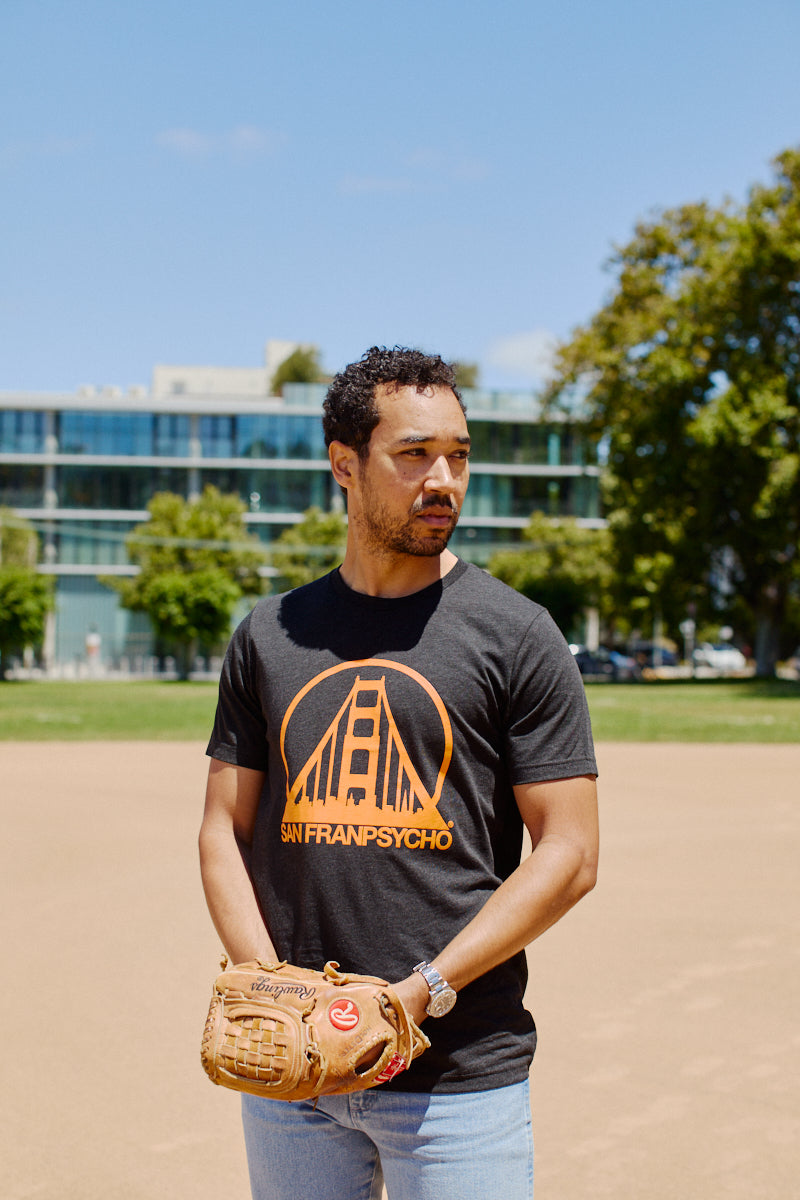 A man holding a baseball glove stands on a field, wearing a super comfortable, hand printed Charcoal & Orange Logo Tee.