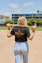 A woman in a Beat LA Black & Orange Tee stands on a baseball field, holding a bat over her shoulders and facing away from the camera.
