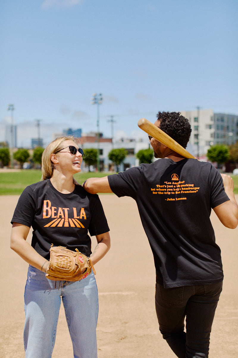 Two people on a baseball field, smiling; one holds a glove, the other rests a bat over their shoulder while wearing the Beat LA Black & Orange Tee.
