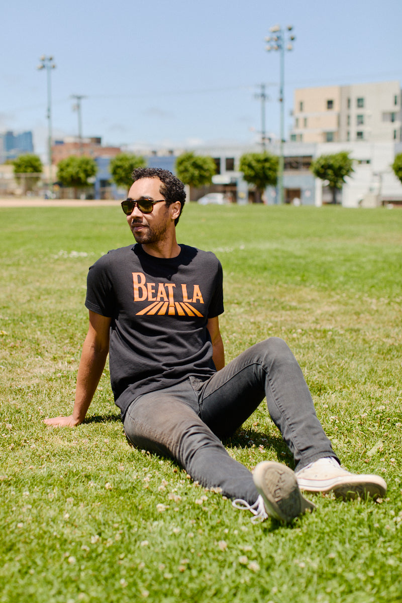 A man in sunglasses and a Beat LA Black & Orange Tee sits on the grass in a sunny park, with city buildings rising in the background.