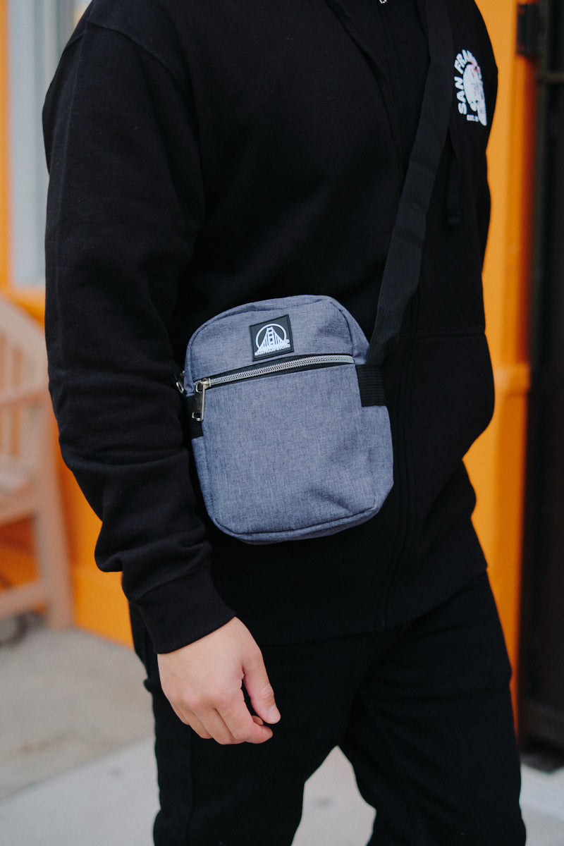 Person in a black hoodie wearing a Small Sling Bag with zippered pockets, standing outside near an orange wall.