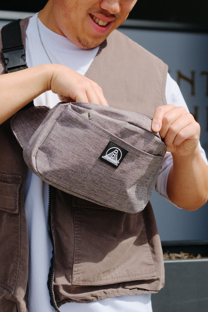 Man smiling and opening a gray Sling Bag with a black patch and zippered pockets, while wearing a brown vest over a white shirt.