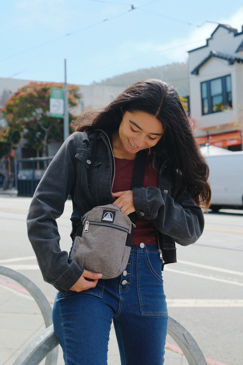 A woman stands on a city street, smiling as she opens her Small Sling Bag featuring an adjustable strap and convenient zippered pockets.