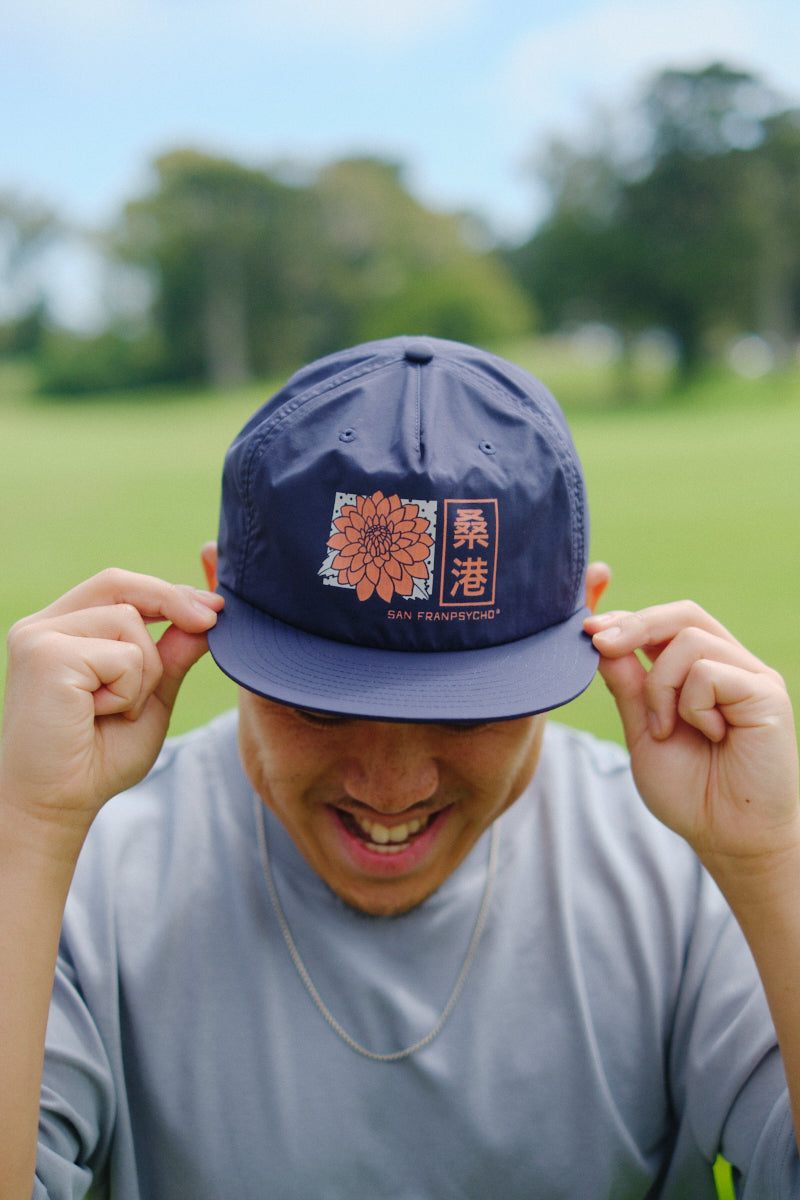 A smiling person adjusts the Sunset Dahlia Hat, a navy blue flat brim cap featuring a flower and Asian characters on the front, while outdoors.
