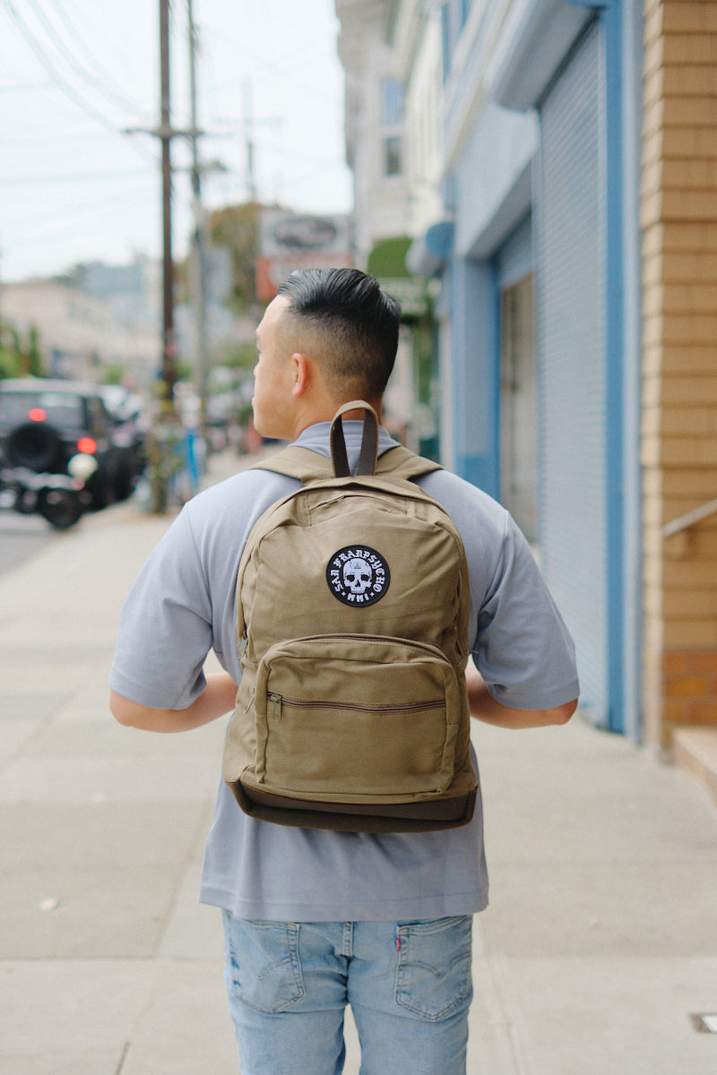 A person carrying the Olive Backpack w/ Punk Skull Patch, detailed with leather trims, walks away on a city sidewalk.