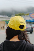 Person with long dark hair wears a classic fit black hoodie and a yellow Smiley Dad Hat, facing away outdoors on a cloudy day.