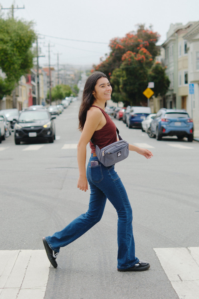 A woman in jeans and a maroon top smiles as she walks across a city crosswalk, carrying the Sling Bag with an adjustable strap stylishly over her shoulder.
