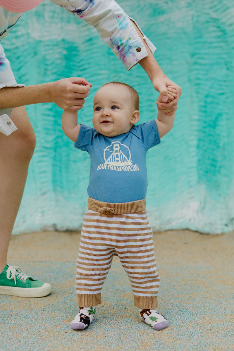 A smiling baby stands with support, wearing the Gull Logo Onesie inspired by Erik Abel and striped pants, set against a teal background.