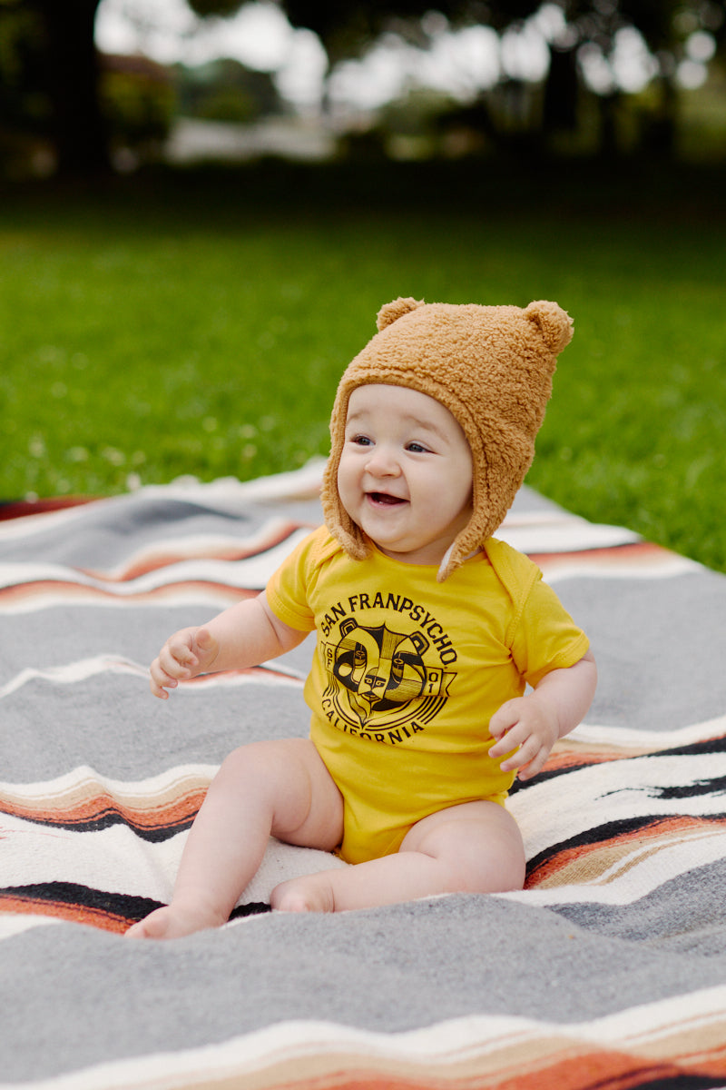 A smiling baby in a Bear Essentials Onesie and bear hat sits on a striped blanket on the grass.