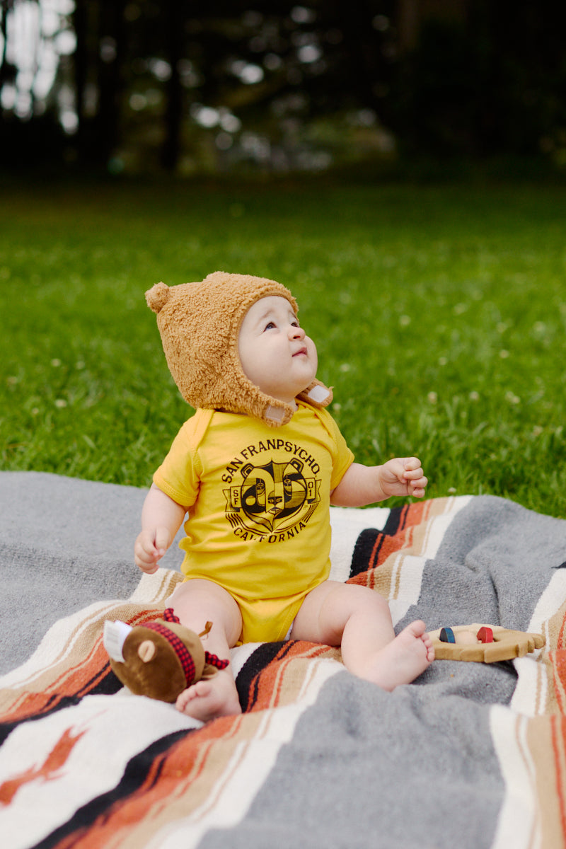 A baby wearing the Bear Essentials Onesie and a bear hat sits on a blanket in the grass—an adorable and practical pick among San Francisco baby clothes.