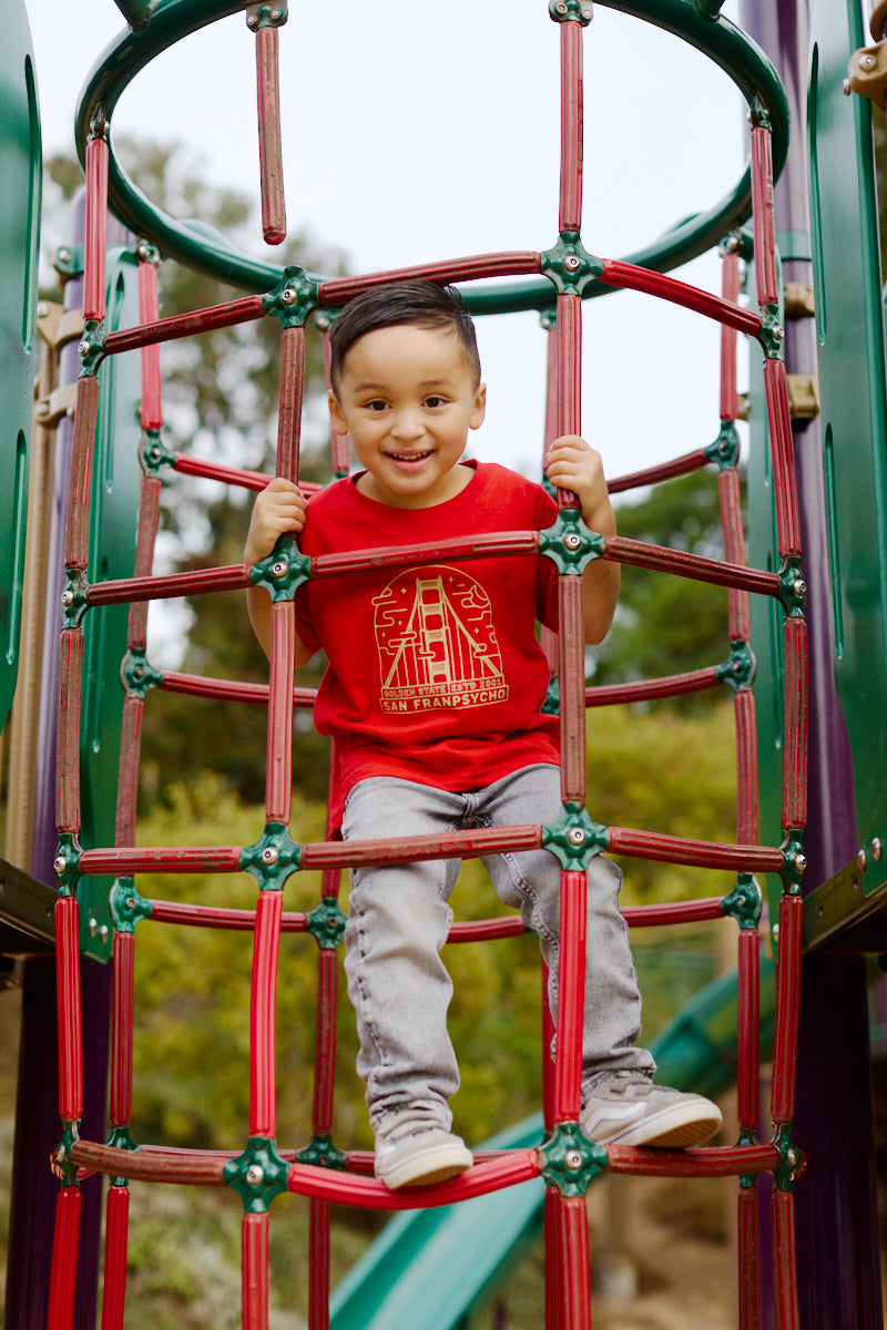 A smiling youth in a red & gold Youth Fogtown Tee climbs on a red and green playground structure outdoors in San Francisco.