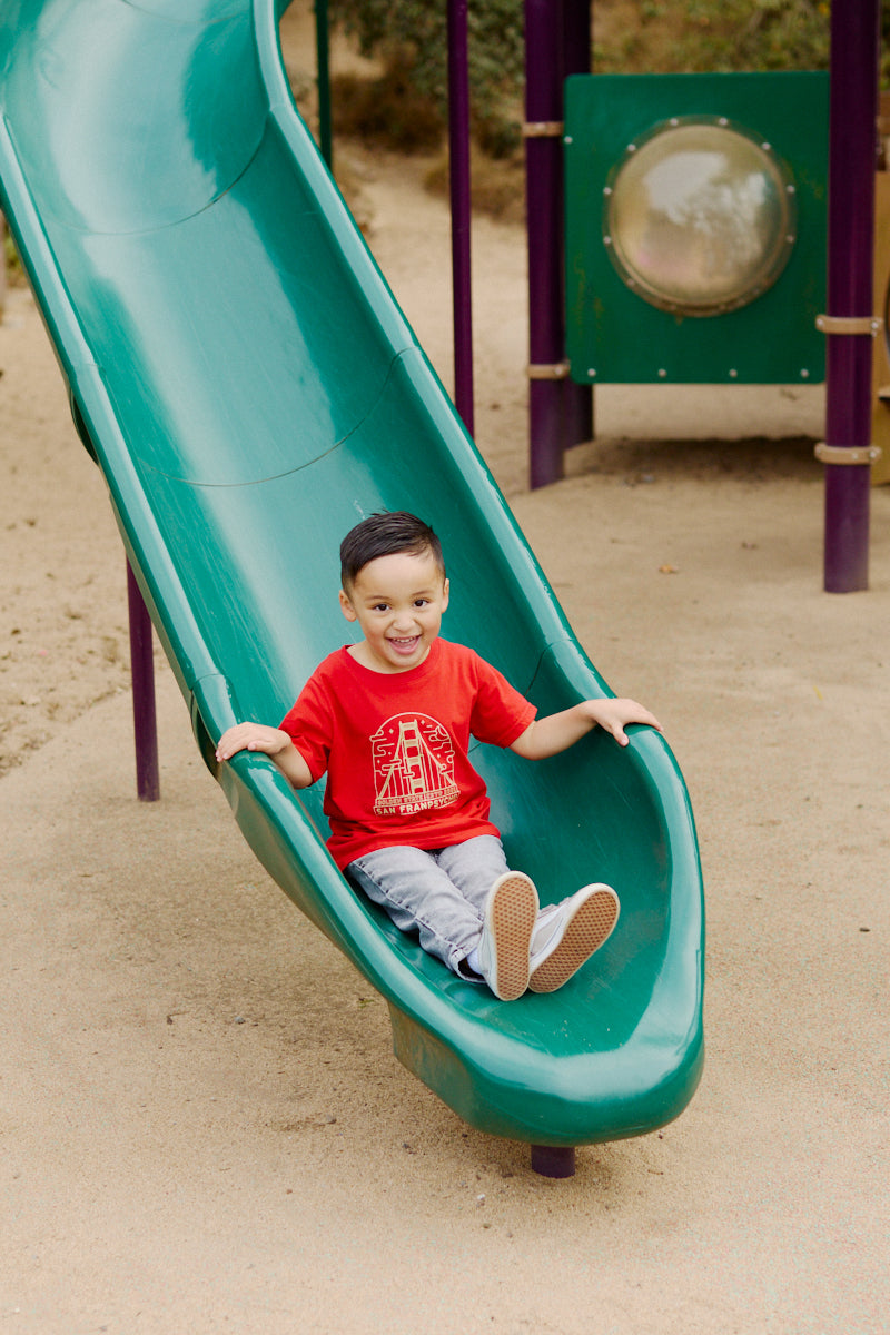 A smiling young boy in a Red & Gold Youth Fogtown Tee slides down a green playground slide onto sandy ground, wearing Fogtown Tees’ San Francisco-inspired design.