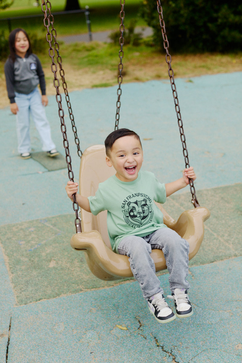 A smiling young boy wearing the Bear Essentials Youth Tee enjoys swinging at the playground, with another child in the background.