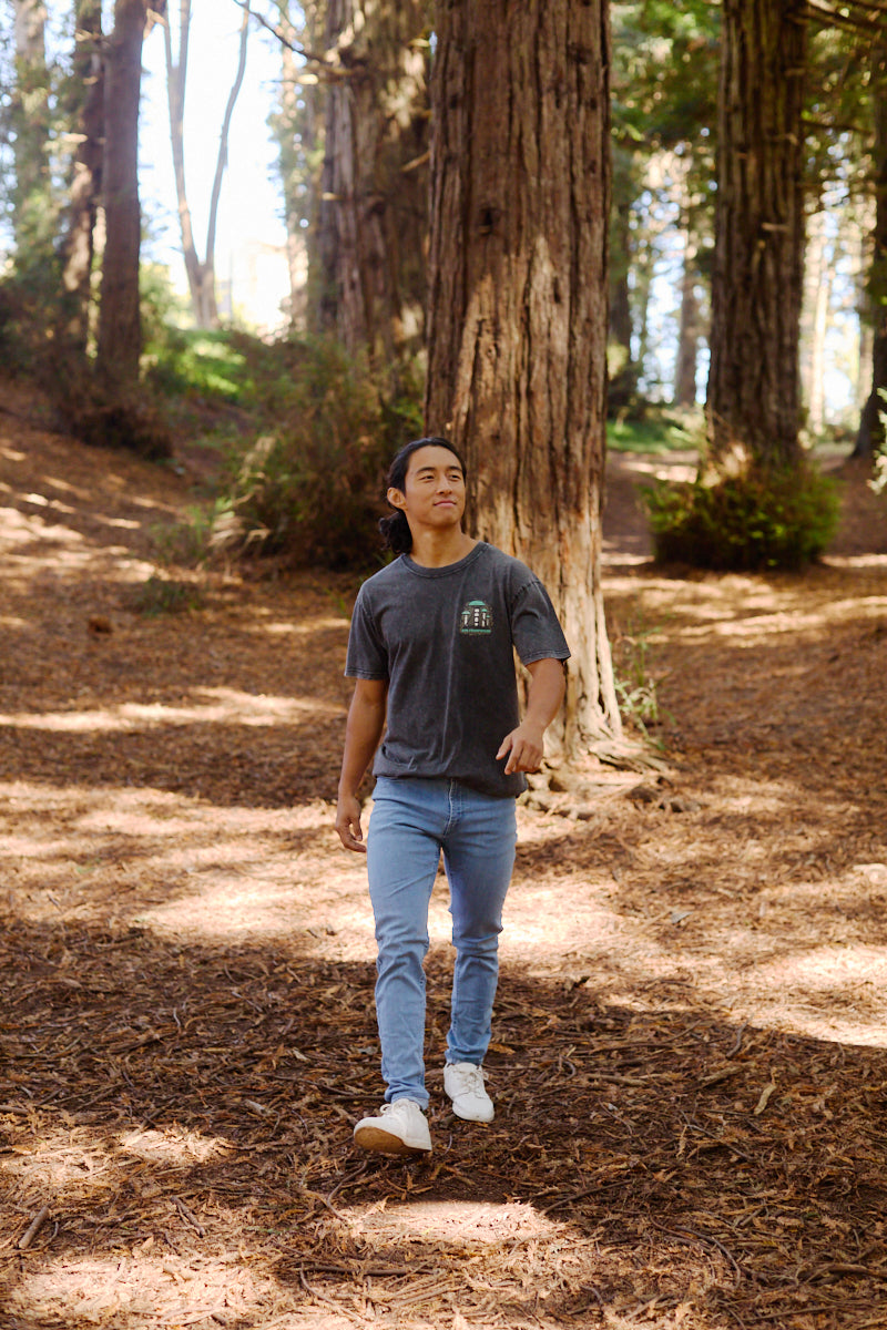 A person strolls through a sunlit forest of tall trees, wearing the Mind Bloom Tee with a skull and mushroom graphic, paired with jeans and white shoes.