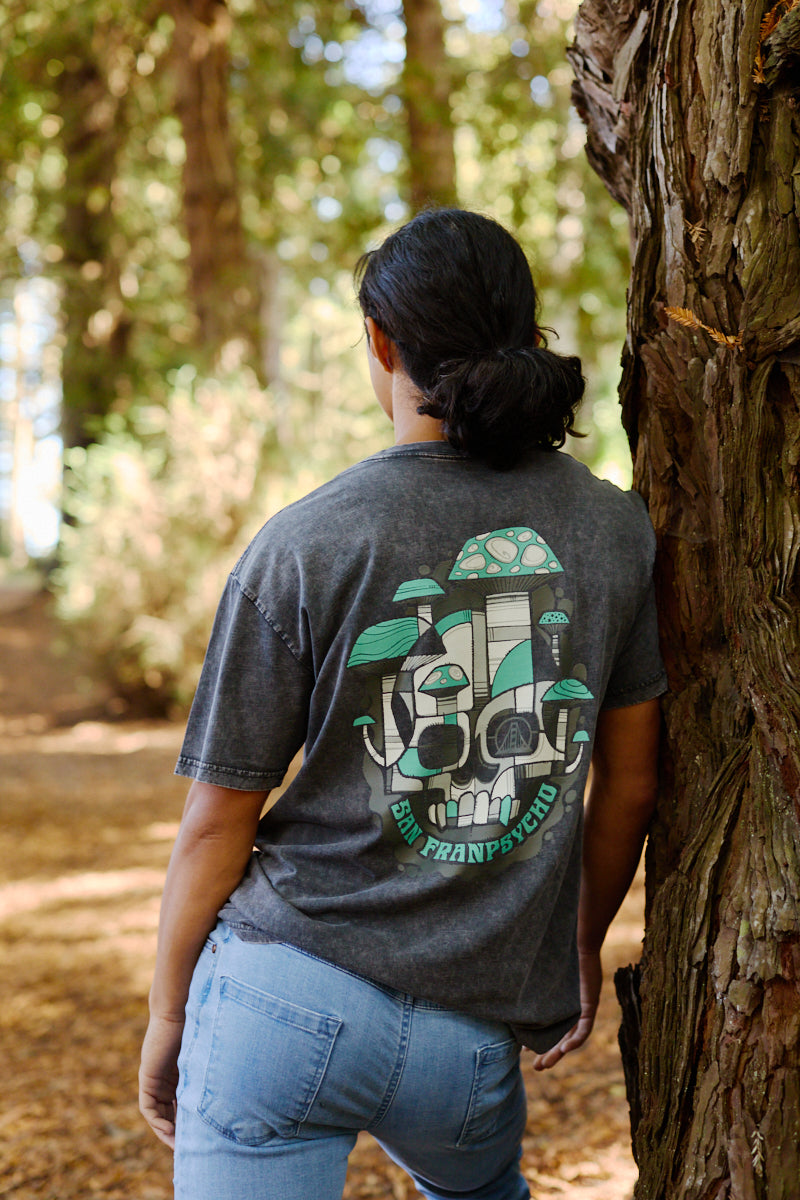 A person in jeans and the Mind Bloom Tee—featuring a skull and mushroom graphic—leans against a tree trunk in a forest, capturing a scene inspired by Erik Abel’s signature style.