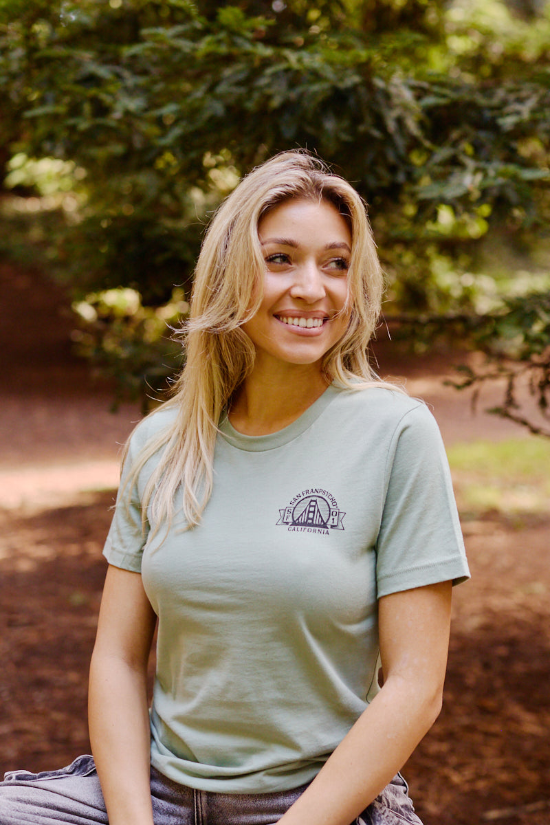 A smiling woman with long blonde hair wears the Bear Essentials Tee in light green, sitting outdoors among trees and capturing a relaxed California bear vibe.