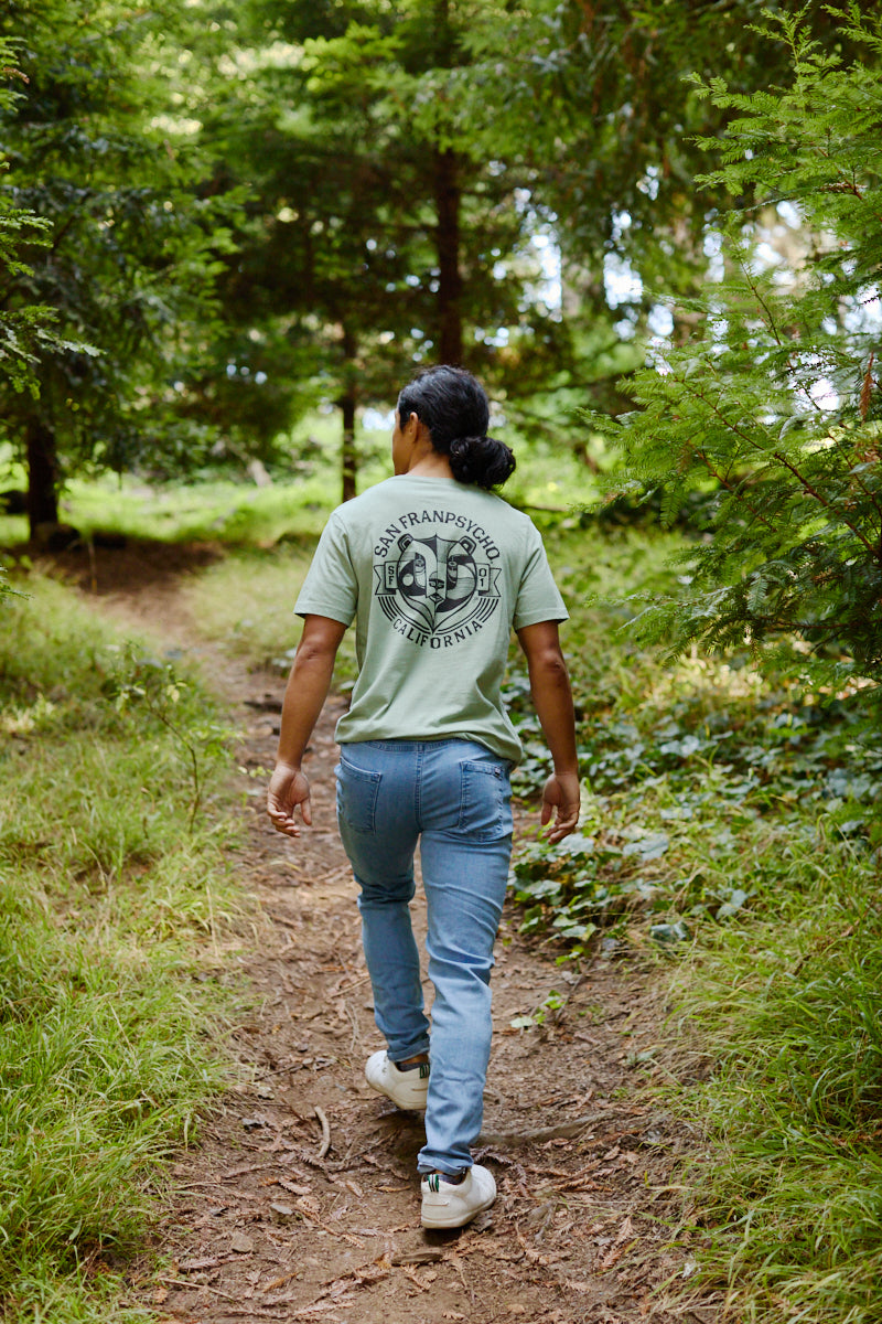 A person walks on a forest trail in jeans and a green Bear Essentials Tee with a California bear and San Francisco graphic on the back.
