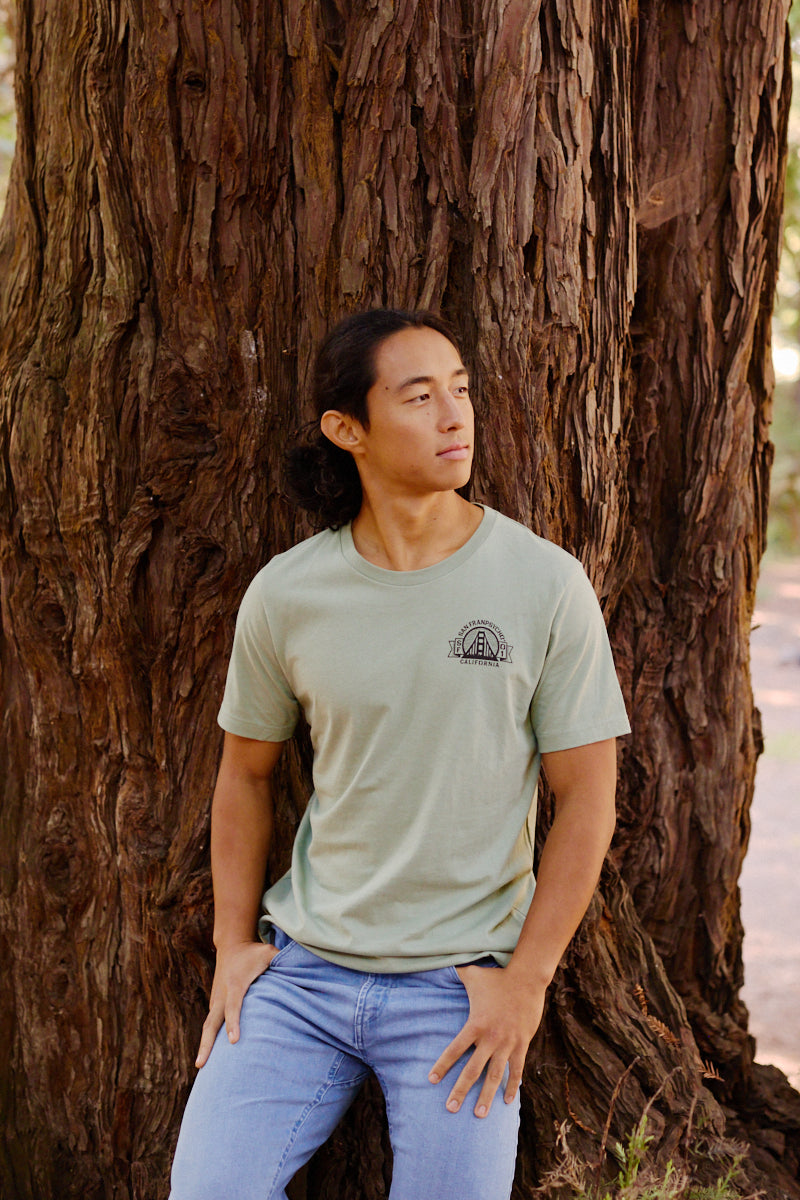 Wearing the Bear Essentials Tee, a man leans against a tree outdoors, gazing to the side and embodying San Francisco's laid-back vibe.