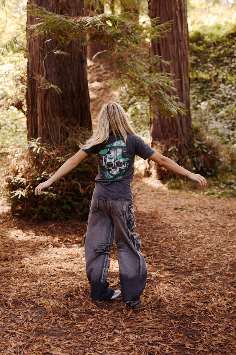 A person with long blond hair and baggy clothes stands with arms outstretched in a forest among tall trees, wearing the Mind Bloom Tee—a mineral wash shirt featuring a skull and mushroom graphic.