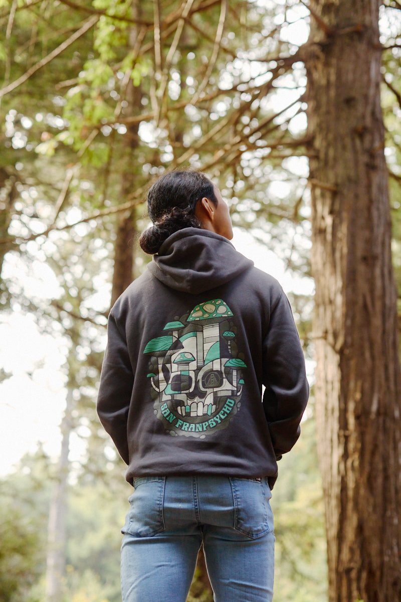 A person wearing the Mind Bloom Pullover Hoodie, with a vibrant skull and mushroom-inspired graphic (in the style of Erik Abel), stands in a forest facing away from the camera, highlighting the hoodie’s unique artistic flair.