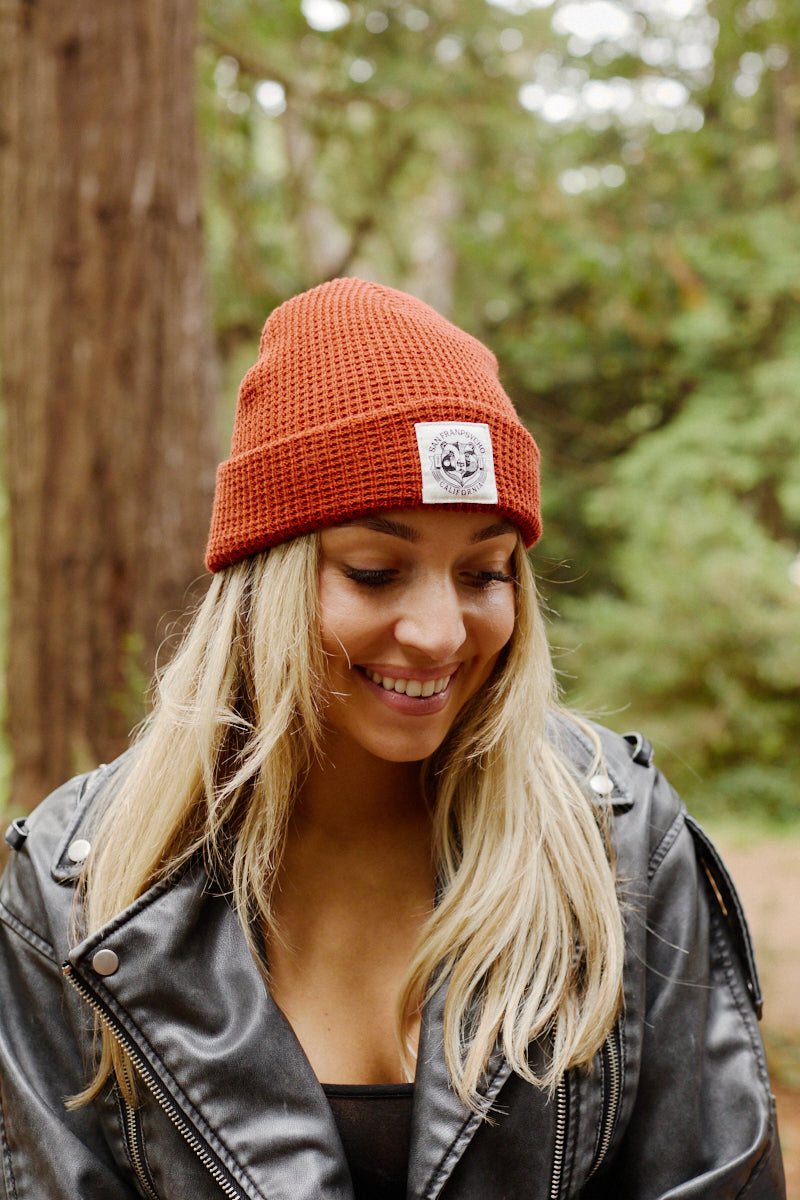 Woman with long blonde hair wearing a Bear Essentials Rust Beanie and black leather jacket, smiling outdoors in a forest.