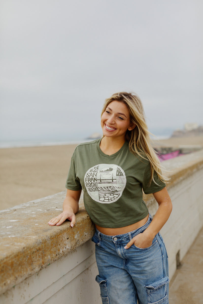 A woman smiles as she leans on a concrete railing at the beach wearing the Women's Elemental Cropped Tee, featuring a green fabric with a San Francisco-inspired graphic design.