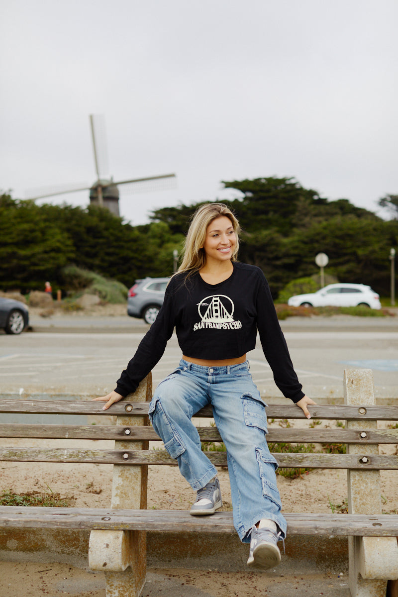 A smiling woman sits on a park bench in front of a windmill, radiating coastal vibes in the Women's Gull Logo Cropped Long Sleeve and cargo jeans.