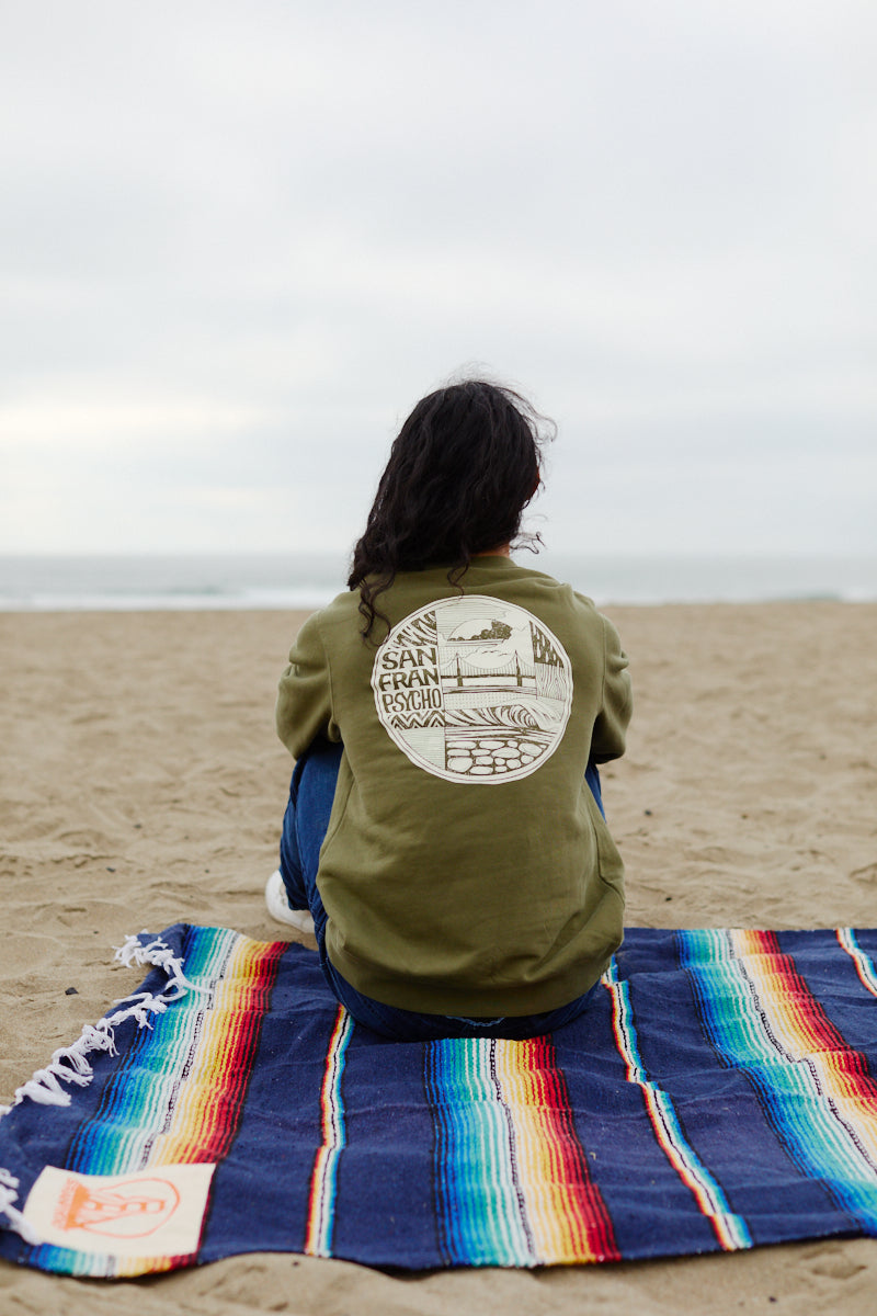 A person sits on a colorful blanket at the beach, facing the ocean, wearing a green Elemental Crewneck made from 100% cotton.