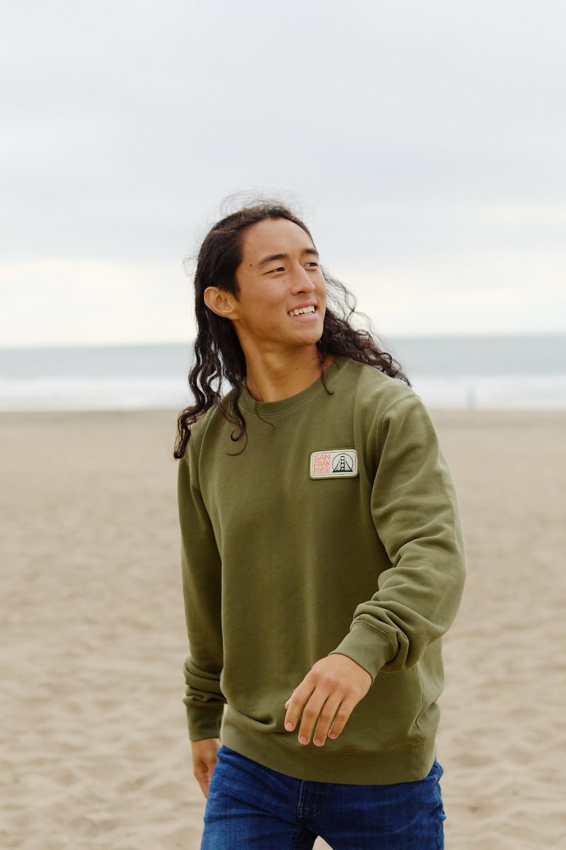 Wearing the Elemental Crewneck in green, a person smiles while walking along a sandy beach with the ocean behind them.
